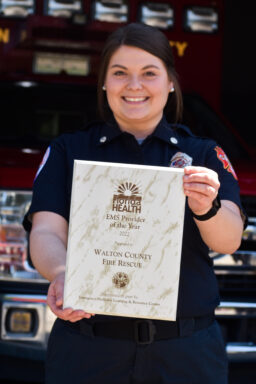 A female EMT holding an award standing in front of a red ambulance. 
