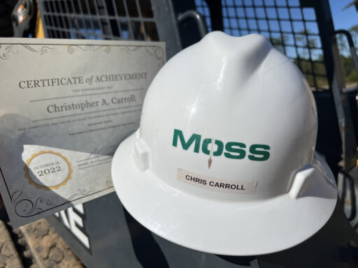A white hard hat reading "Moss" with the name Christopher Carroll on the front sits beside a Certificate of Achievement